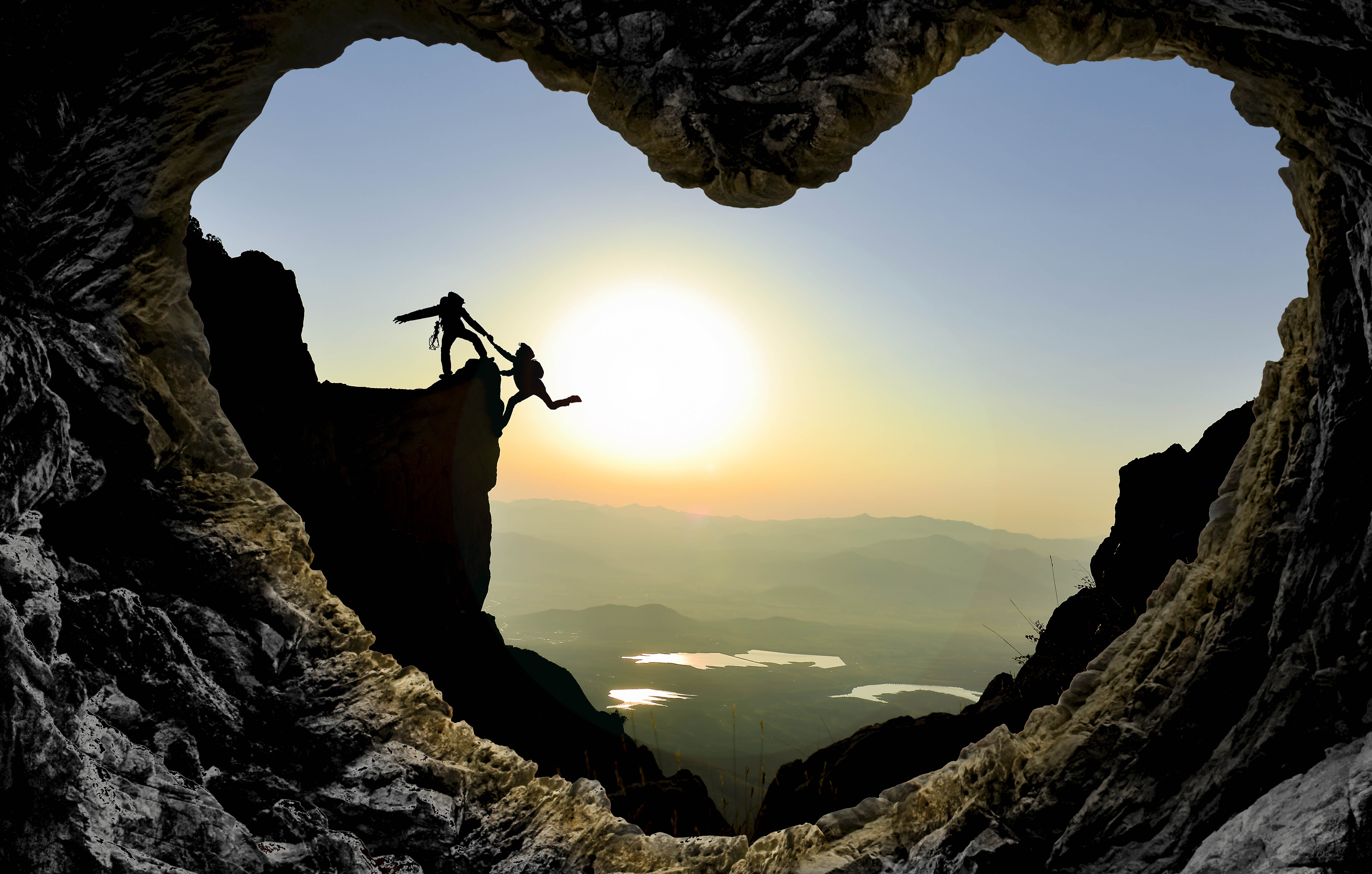 Cave in the shape of a heart, with the silhouette of two people - one helping the other up the cliff - in the twilight